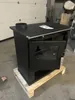 A black wood-burning stove is shown on a wooden pallet in what appears to be a warehouse or workshop. Produktbild-Vorschau