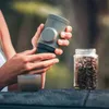 A person is holding a portable espresso maker next to a jar of coffee beans and a small cup. Produktbild-Vorschau