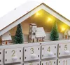 A close-up view of a white wooden advent calendar shaped like a house, decorated with miniature Christmas trees and a church with a clock tower. Warm LED lights are visible inside the roof. Voir les images du produit