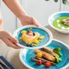 A person is holding a plate with a dessert and other plates with food are visible in the background. Náhled obrázku produktu