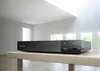 A black Sony Blu-ray player is placed on a light-colored wooden table in a bright, modern room with large windows and a skylight. Voir les images du produit