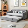 A grey sofa bed with an extended trundle bed, set in a living room with artwork and a wooden cabinet. Produktbild-Vorschau