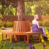 A woman sits on a circular wooden bench around a tree in a garden. Produktbild-Vorschau