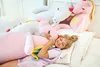 A young girl is sleeping on a bed, hugging a large pink plush unicorn. Other plush unicorns are visible in the background. Produktbild-Vorschau