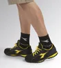 A person's legs wearing black and yellow Diadora safety shoes and black socks with a white logo. Náhľad obrázku produktu