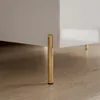 A close-up shot of the leg of a white, high-gloss coffee table with gold-colored legs, resting on a wooden floor. Náhled obrázku produktu