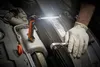 A person wearing gloves is working on a car engine, illuminated by a portable work light. Produktbild-Vorschau