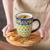 Zwei Hände halten einen bunten Keramikbecher mit Tee und Zitrone auf einem Holztablett. Náhled obrázku produktu
