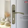 A close-up of a bronze door handle on a white door, with a blurred interior scene in the background. Produktbild-Vorschau