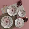 A top-down view of a set of dinner plates with a rustic, abstract design, arranged on a pink tablecloth with grapes and forks. Produktbild-Vorschau