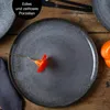 A close-up shot of a dark gray dinner plate with a small orange flower on it, set on a wooden table. Produktbild-Vorschau