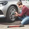 A man is kneeling on a paved surface, using a cordless impact wrench to tighten the lug nuts on a car wheel. A torque wrench is lying on the ground in the foreground. Podgląd zdjęcia produktu