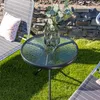 A small round glass table with a vase of greenery on it, placed on a lawn next to a lounge chair. Produktbild-Vorschau