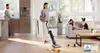 A woman is using a wet dry vacuum cleaner to clean up a spill on a wooden floor in a modern living room. A man is in the background in a kitchen area, and a child and dog are on a sofa. Náhled obrázku produktu