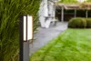 A modern outdoor path light illuminates a garden path next to lush green grass and foliage. Anteprima dell’immagine prodotto