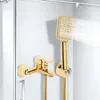 A gold-colored shower faucet and handheld showerhead are mounted on a white wall, with water spraying from the showerhead. Podgląd zdjęcia produktu