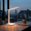 A desk lamp is on a wooden table next to a laptop and a mug, with a city view in the background. Produktbild-Vorschau