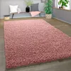 A shaggy pink rug is laid out on a wooden floor in a living room setting. A bench with pillows and a plant are visible in the background. Produktbild-Vorschau
