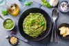 A close-up overhead shot of a bowl of spaghetti with pesto sauce, surrounded by ingredients like pine nuts, garlic, olive oil, and basil. Produktbild-Vorschau