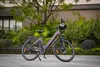 An electric bicycle is parked on a paved surface in front of a building with greenery. Produktbild-Vorschau