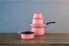 A stack of pink pots and pans with a single pot in the foreground, all on a wooden table against a gray background. Anteprima dell’immagine prodotto