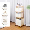 A bamboo laundry cart with three fabric baskets is shown in a room with white curtains in the background. The cart has wheels and is made of natural bamboo. The baskets are beige and have handles. The top shelf has two bottles of soap and a diffuser. Anteprima dell’immagine prodotto