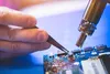 A close-up shot of a person's hand using tweezers to manipulate a component on a blue circuit board, with a hot air soldering station in the background. Produktbild-Vorschau