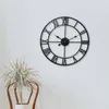 A black analog wall clock with Roman numerals on a white wall, with a potted plant in a chair in the foreground. Voir les images du produit