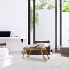 A modern living room with a laptop on a bamboo table in the foreground, and a large window with greenery outside. Produktbild-Vorschau