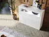 A white, high-gloss sideboard with LED lighting is shown in a modern living room setting, placed on a herringbone wooden floor with a rug in the foreground. Podgląd zdjęcia produktu