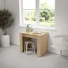 A compact wooden table with a stool tucked underneath, placed next to a window with greenery outside. A chair is positioned to the right. Anteprima dell’immagine prodotto