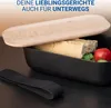 A black lunchbox with a wooden lid, containing a baguette, cucumber slices, and tomatoes, sits on a dark surface with a black elastic strap next to it. Text is overlaid at the top. Produktbild-Vorschau