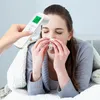 A woman is lying in bed, wrapped in a blanket, and blowing her nose into a tissue while a non-contact infrared thermometer is held to her forehead. Produktbild-Vorschau