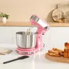 A pink stand mixer with a stainless steel bowl is shown on a white countertop, with sliced bread and a spatula nearby. The background features a kitchen counter with a plant and some jars. Produktbild-Vorschau
