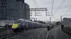 A purple and yellow train is traveling on train tracks in a city on a rainy day. Náhled obrázku produktu