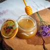 A jar of honey with a honey dipper, on a wooden surface with flowers. Náhľad obrázku produktu