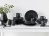 A collection of black stoneware dinnerware, including plates, bowls, and mugs, arranged on a white surface with a plant in a vase on the left. Produktbild-Vorschau
