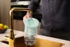 A person is filling a water filter pitcher under a golden faucet in a kitchen sink. Náhľad obrázku produktu