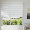 A white zebra roller blind is installed on a window, with a view of a green landscape and flowers outside. Voir les images du produit