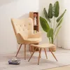 A beige armchair and ottoman are placed on a rug in a room with a plant and a small bookshelf. Náhled obrázku produktu