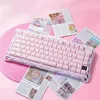 A pink mechanical keyboard is displayed on a pink surface, surrounded by various printed materials that appear to be magazine pages or promotional flyers. Produktbild-Vorschau