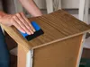A person is applying a wood-grain adhesive film to a piece of furniture with a blue applicator. Produktbild-Vorschau