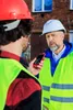 Two construction workers in hard hats and high-visibility vests are talking, with one holding a red smartphone. Podgląd zdjęcia produktu