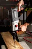 A person is using a hammer drill to drill into a wooden plank in what appears to be a workshop or garage setting. Podgląd zdjęcia produktu