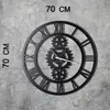 A large, black, metal wall clock with Roman numerals and gears, mounted on a concrete wall. Text indicating the size is present. Náhľad obrázku produktu