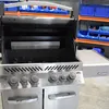A close-up view of a stainless steel grill with its lid open, showing the burners and a warming rack. Shelving units with blue bins are visible in the background. Produktbild-Vorschau