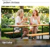 Two women are enjoying a meal and drinks at a picnic table in a garden setting. Produktbild-Vorschau
