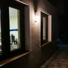 An outdoor wall lamp illuminates a textured wall with two windows at night. Produktbild-Vorschau