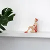 A decorative figurine of a woman in a vintage swimsuit sitting on a shelf next to a large green leaf. Voir les images du produit