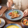 A person is eating a meal from a blue plate with shrimp, vegetables, and potatoes. There are other dishes and cutlery on a wooden table. Náhled obrázku produktu